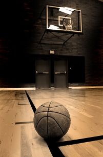 fototapeta z leżącą na pierwszym planie piłką do gry w koszykówkę leżącą na parkiecie a w tle nad czarnymi dzwiami widać obręcz kosza