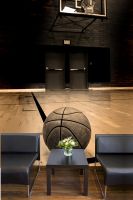 fototapeta z piłką do koszykówki na tle hali sportowej za dwoma czarnymi fotelami i stolikiem z bukietem kwiatów