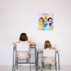Obraz na płótnie przedstawiający księżniczki Disneya powieszony nad stolikami przy których siedzą dwie dziewczynki