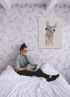 Obraz z wizerunkiem lamy powieszony w pokoju na kwiecistej różowej ścianie nad łóżkiem z dziewczyną obsługującą laptopa