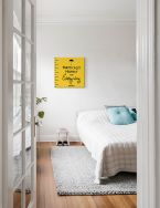 Żółty obraz z napisem Practically Perfect z filmu Mary Poppins zawieszony w sypialni nad łóżkiem z białą pościelą