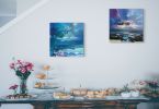 Obraz z widokiem na fioletowe wybrzeże i góry powieszony w salonie nad stołem z jedzeniem i kwiatami