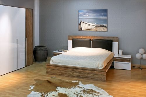 aranżacja obrazu na płótnie przedstawiającego białą łódź zacumowaną na piaszczystej plaży na tle spokojnego morza i zachmurzonego nieba w sypialni z szarymi ścianami nad dużym drewnianym łóżkiem