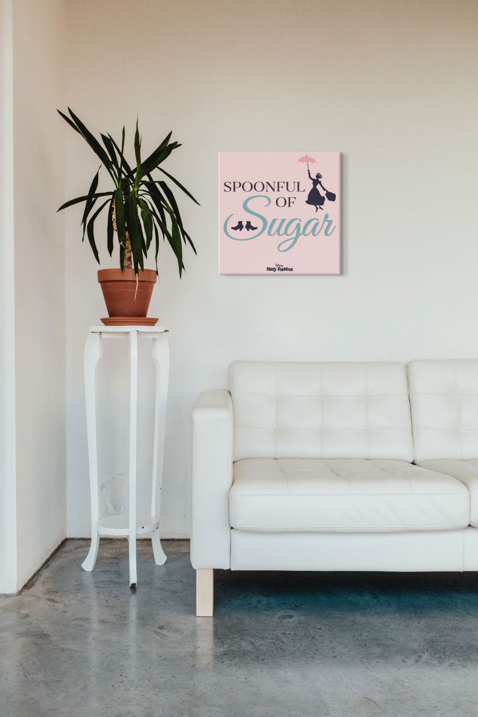 Różowy obraz z napisem Spoonful of Sugar z filmu Mary Poppins powieszony w salonie nad skórzaną białą kanapą obok kwiatka w doniczce