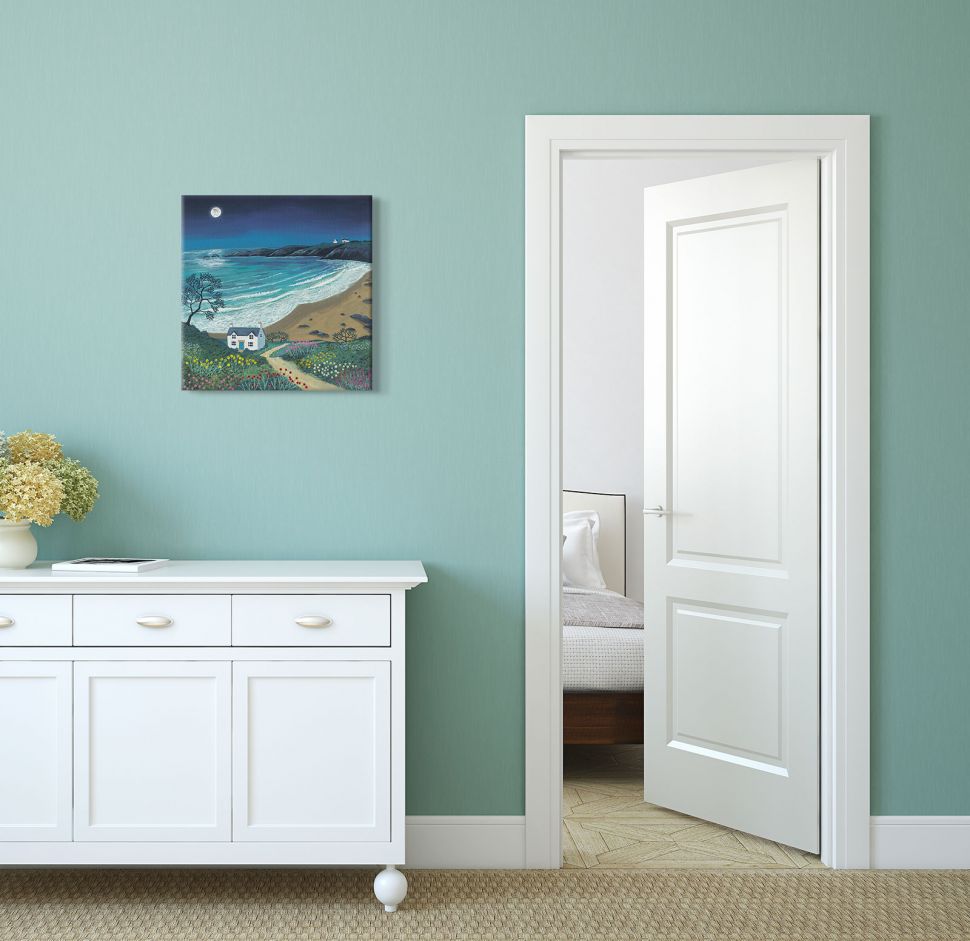 Obraz z domem, plażą i morzem wieczorną porą powieszony w pokoju na niebieskiej ścianie nad białą szafką z kwiatami